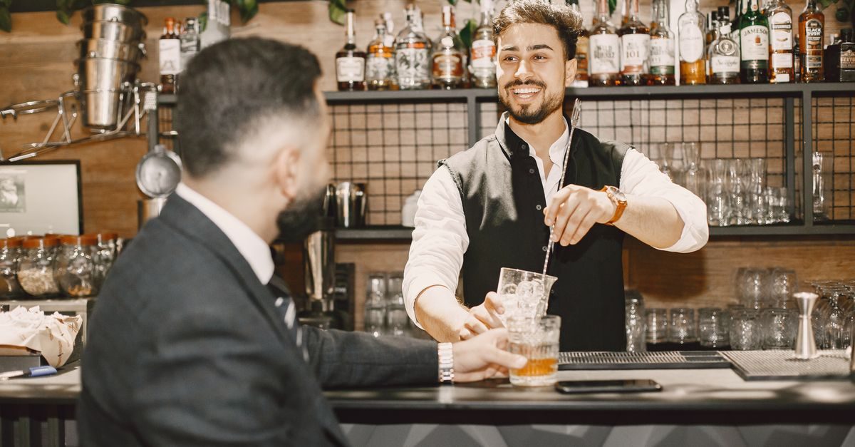 Camarero sonriente prepara una bebida para un cliente en la barra de un restaurante con estanterías de licores al fondo