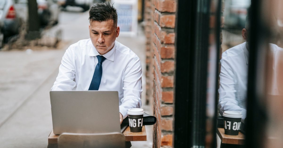 Man in wit overhemd met das werkt geconcentreerd op zijn laptop aan een tafeltje op een stadsstoep met een koffiebeker