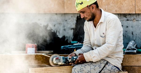 A focused worker wearing a yellow cap uses an angle grinder to cut stone, creating a cloud of dust at a construction site