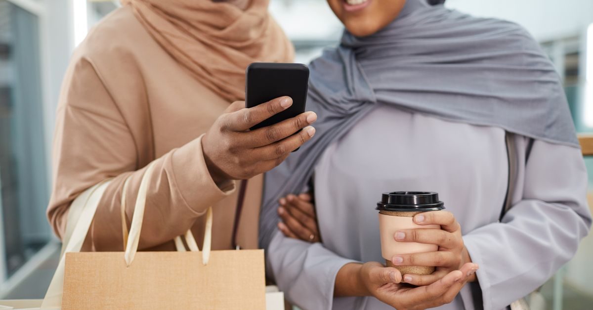 Deux femmes en hijab souriantes regardent un smartphone en tenant un sac de shopping et un gobelet de café dans un centre