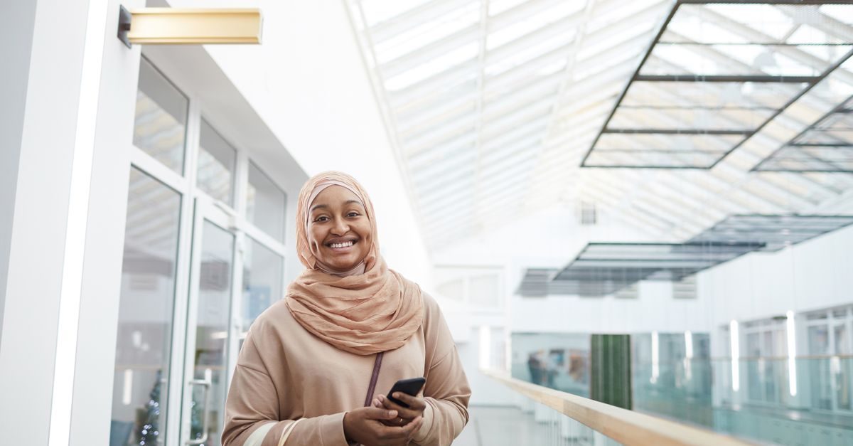 A smiling woman in a beige hijab holds a smartphone while standing in a bright, modern office hallway with glass walls
