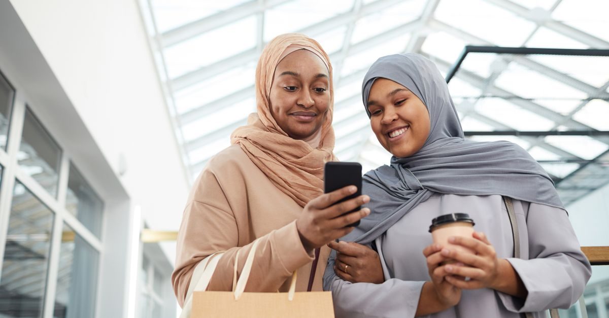 Deux femmes en hijab sourient en regardant ensemble un smartphone dans un centre commercial lumineux