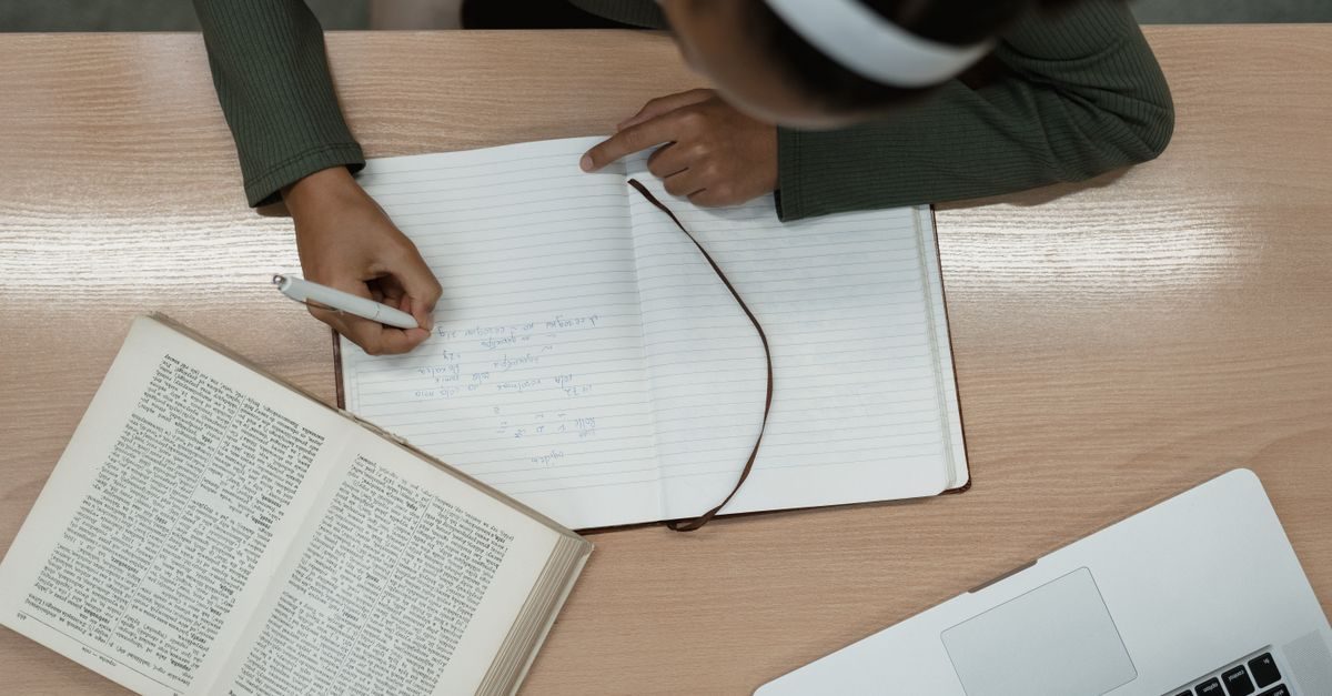 Top-down view of a person writing in a notebook at a desk with an open book and a laptop nearby