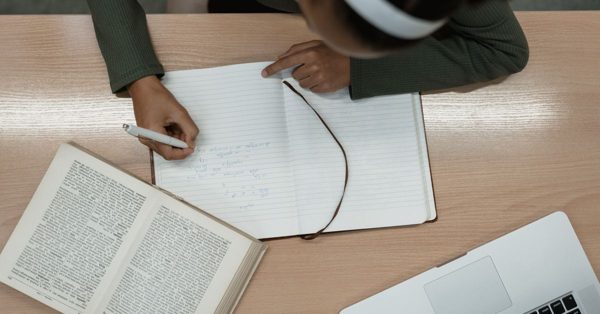 Top-down view of a person writing in a notebook at a desk with an open book and a laptop nearby