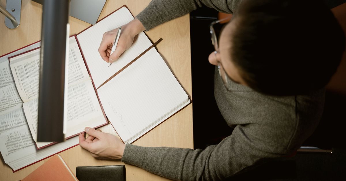 A person wearing glasses sits at a desk, focused on writing in an open notebook next to a large reference book