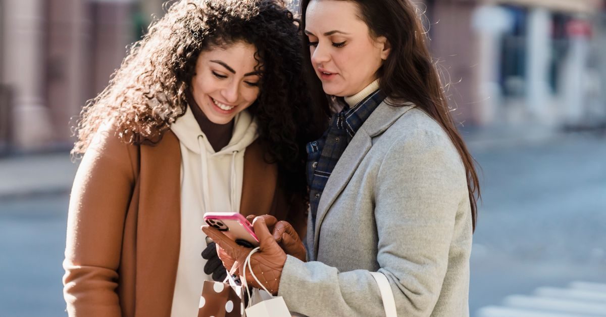 Twee vrouwen kijken glimlachend naar een smartphone terwijl ze met boodschappentassen op een stadsstraat staan