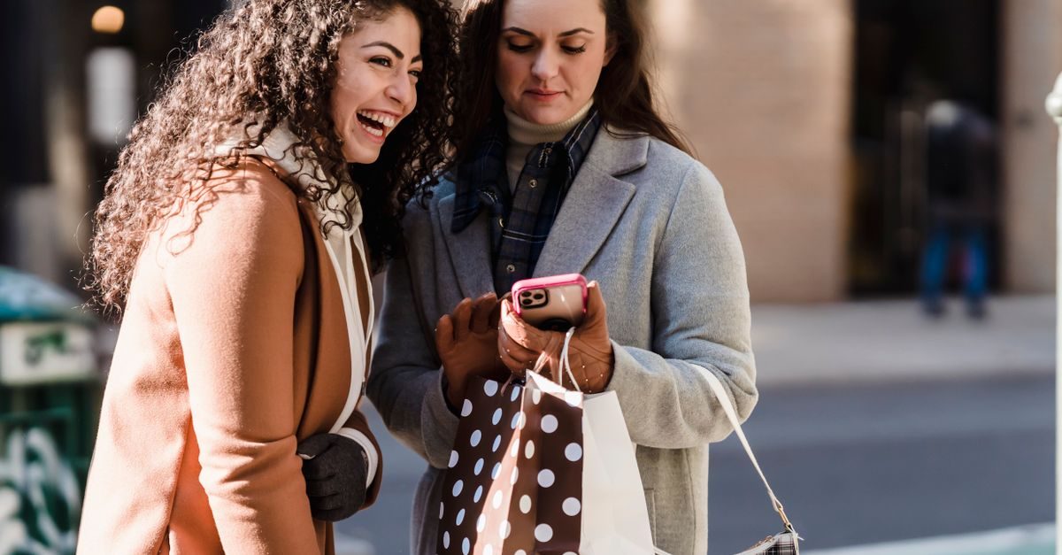 Two women with shopping bags stand on a city street, one laughing while looking at a smartphone held by the other