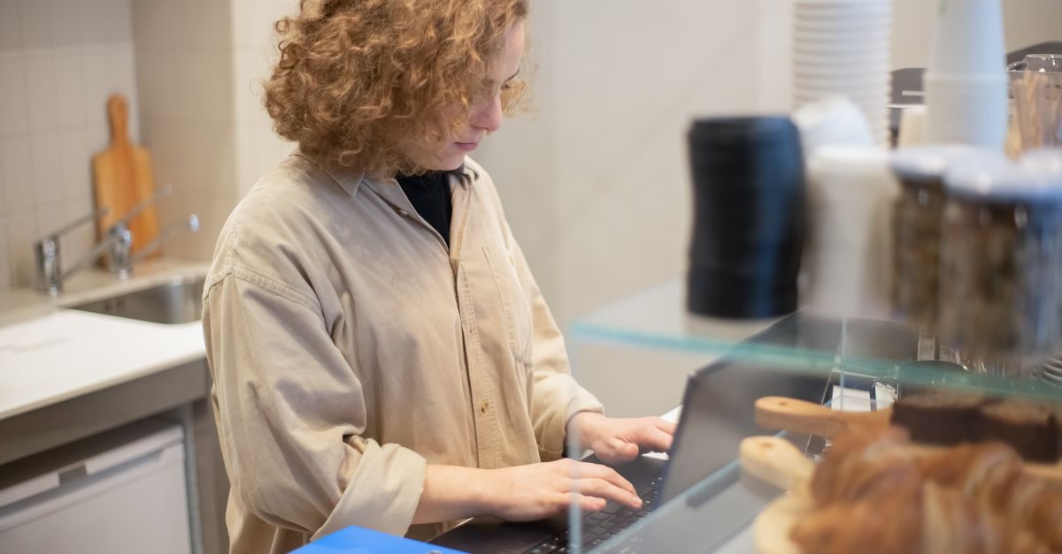 Eine junge Frau mit lockigem Haar arbeitet konzentriert an einem Laptop hinter einer Theke in einem Café