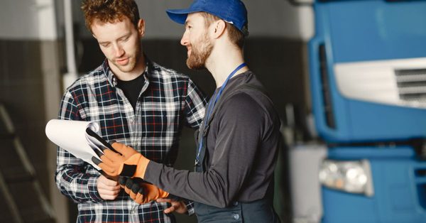 Monteur in werkkleding en een man in ruitjeshemd bespreken samen documenten in een garage met een blauwe vrachtwagen