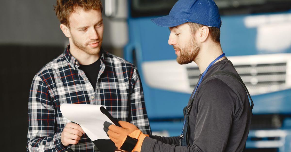 Mechanik w czapce i rękawicach rozmawia z klientem w warsztacie, wskazując na dokumenty trzymane przez mężczyznę w koszuli