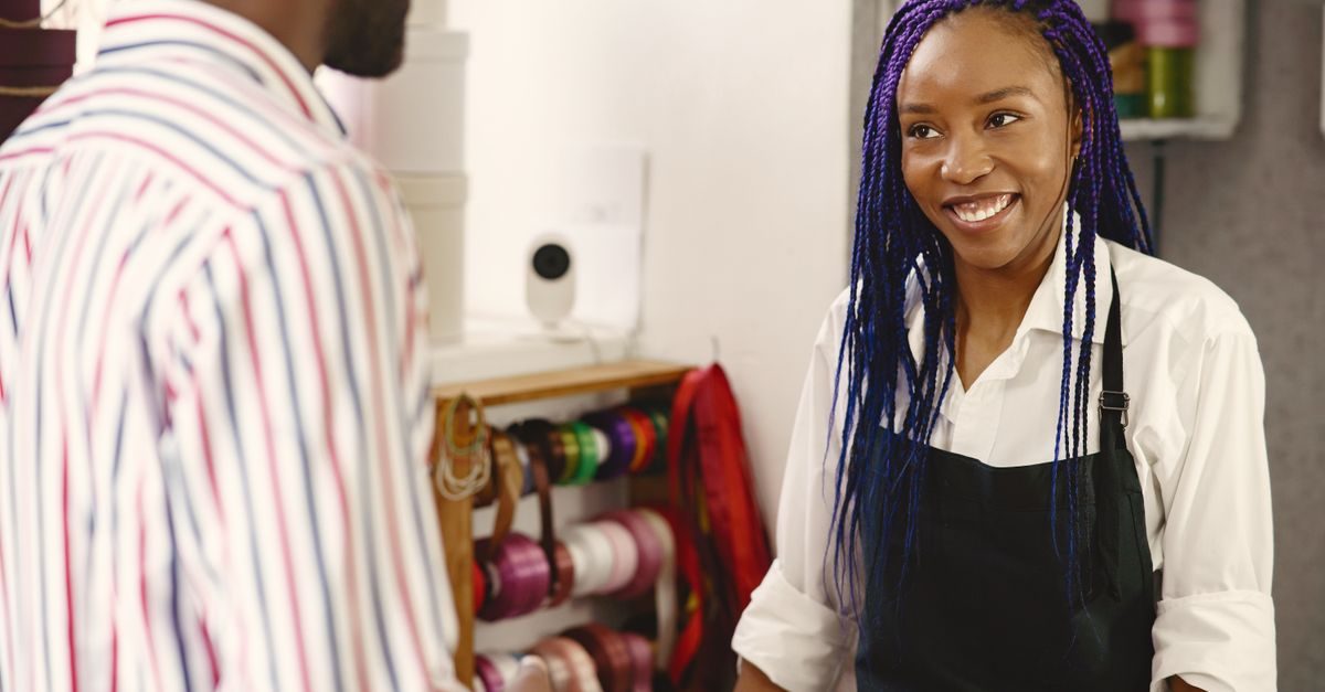 Dependienta con trenzas azules sonríe a un cliente en una tienda con estanterías de cintas de colores al fondo
