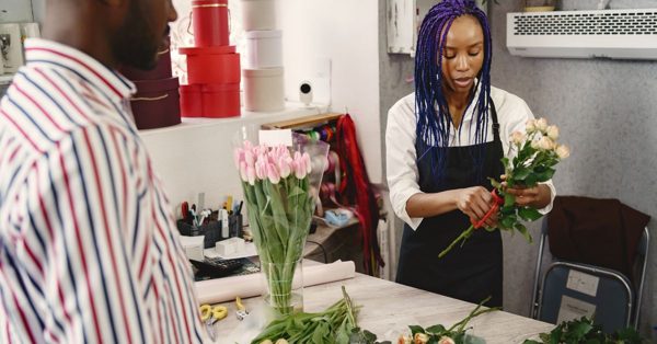 Florista con trenzas azules corta tallos de rosas en su tienda mientras atiende a un cliente frente al mostrador