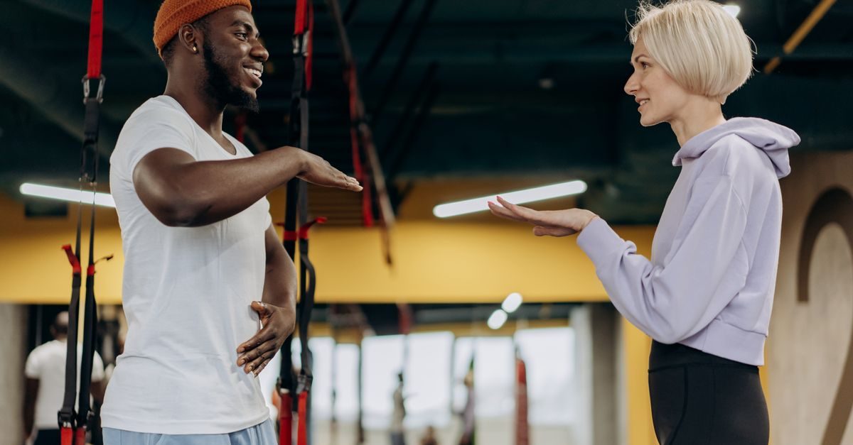 Un homme et une femme souriants discutent dans une salle de sport devant des sangles de suspension TRX