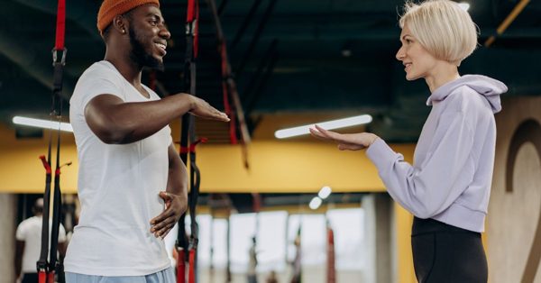 Un homme et une femme souriants discutent dans une salle de sport devant des sangles de suspension TRX