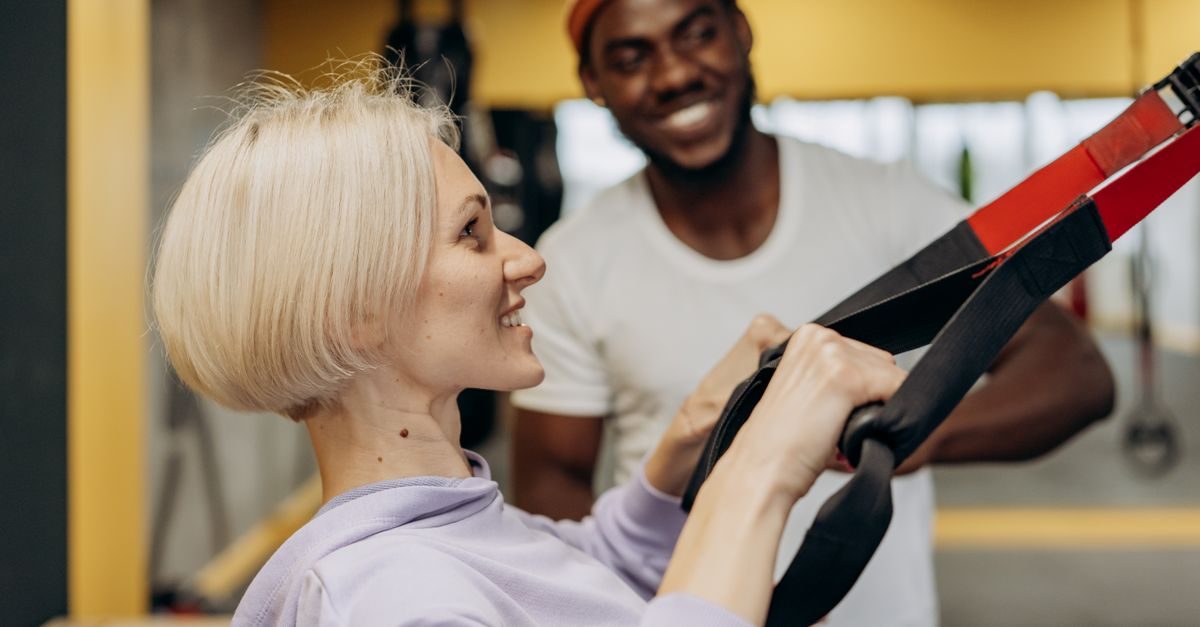 Une femme blonde s'entraîne avec des sangles TRX sous le regard bienveillant et souriant d'un coach sportif en salle