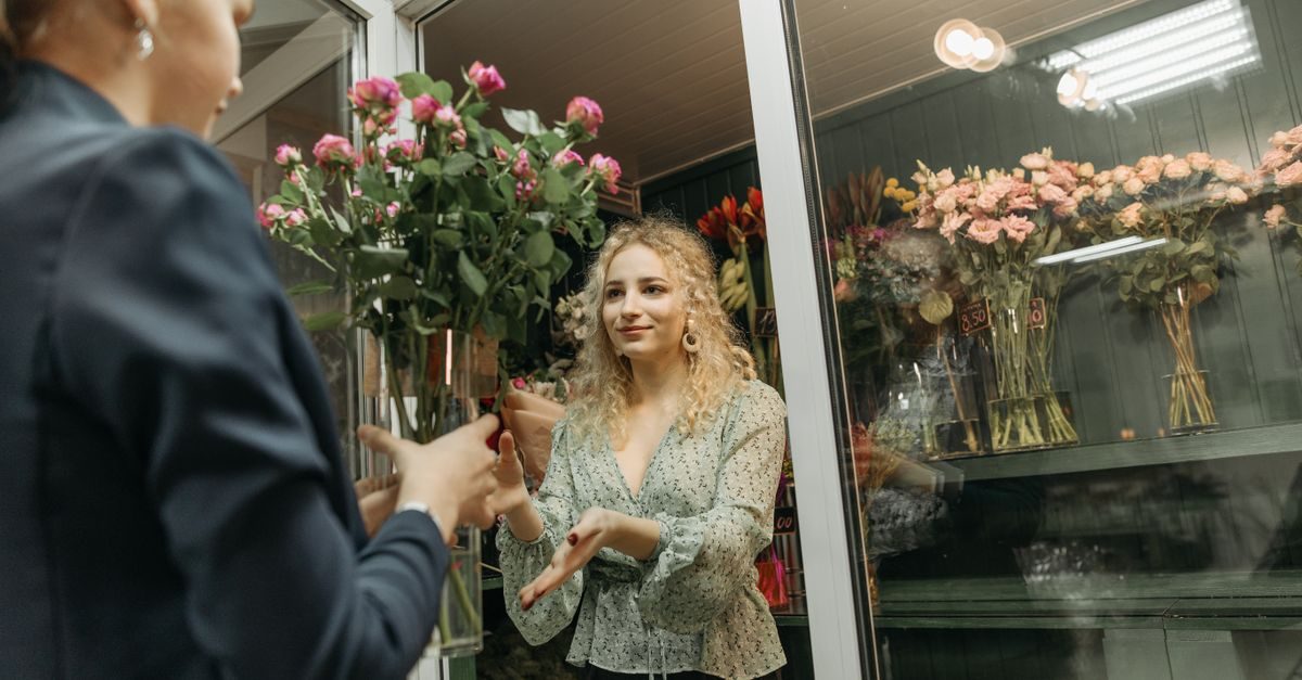 A florist with curly blonde hair smiles while handing a large bouquet of pink roses to a customer in a flower shop