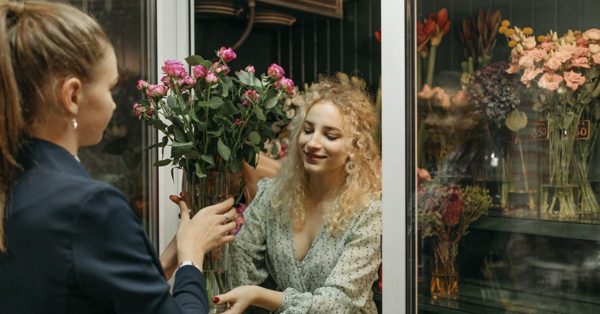 Uśmiechnięta florystka podaje klientce wazon z różowymi różami wewnątrz kwiaciarni z przeszkloną chłodnią pełną kwiatów