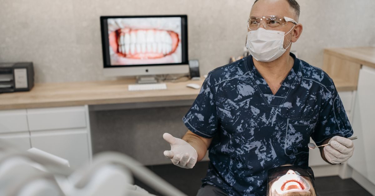 Dentist in scrubs and protective gear consults with a patient in a modern clinic with a dental X-ray displayed on a monitor