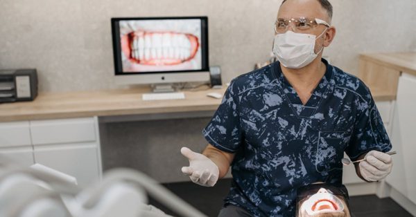 Dentist in scrubs and protective gear consults with a patient in a modern clinic with a dental X-ray displayed on a monitor