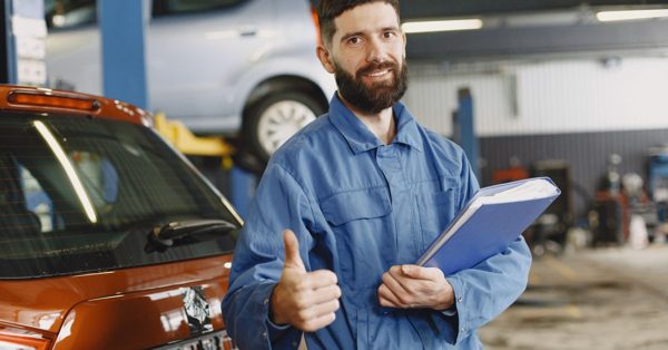 Uśmiechnięty mechanik w niebieskim kombinezonie trzyma dokumenty i pokazuje kciuk w górę w warsztacie samochodowym