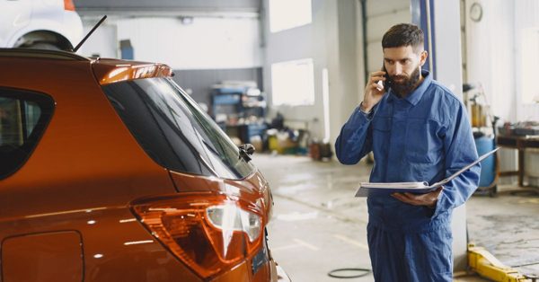 Ein Mechaniker in blauer Arbeitskleidung telefoniert in einer Autowerkstatt mit einem Klemmbrett in der Hand