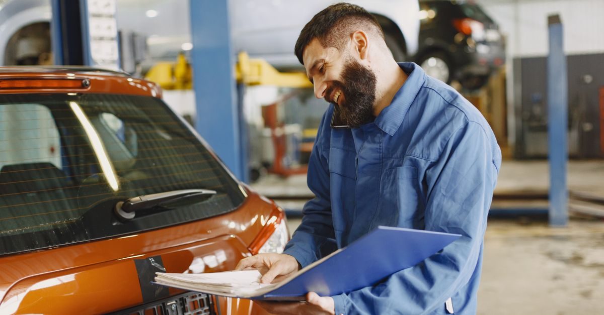 Lachende automonteur in blauwe overall belt met een map in zijn hand bij een auto in een garage