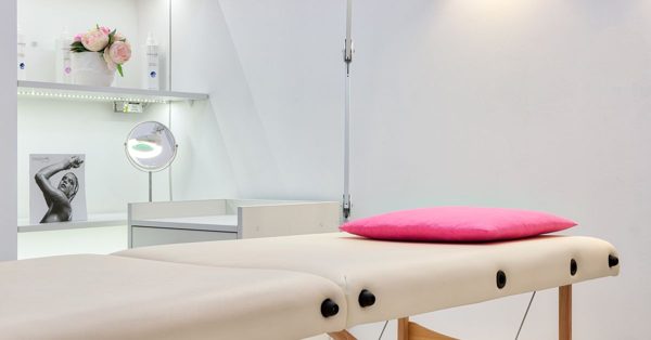 Camilla de masaje blanca con cojín rosa en una sala luminosa y minimalista con estantes decorados y un espejo de aumento