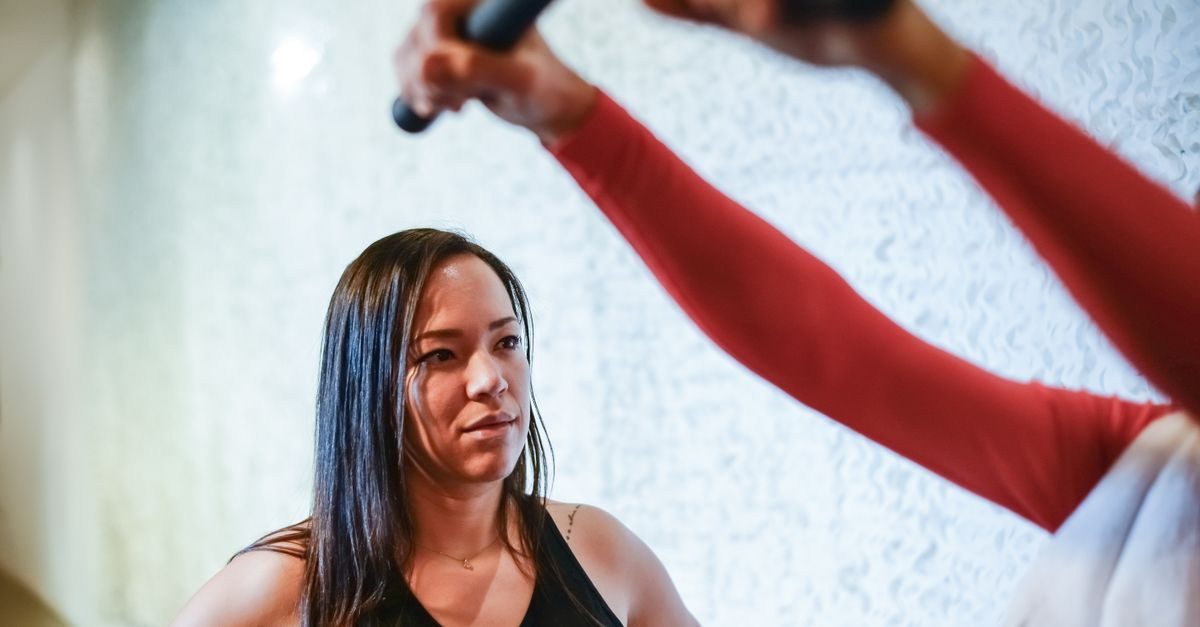 A focused woman watches a person in a red long-sleeved shirt lifting a black exercise bar in a bright, modern gym studio