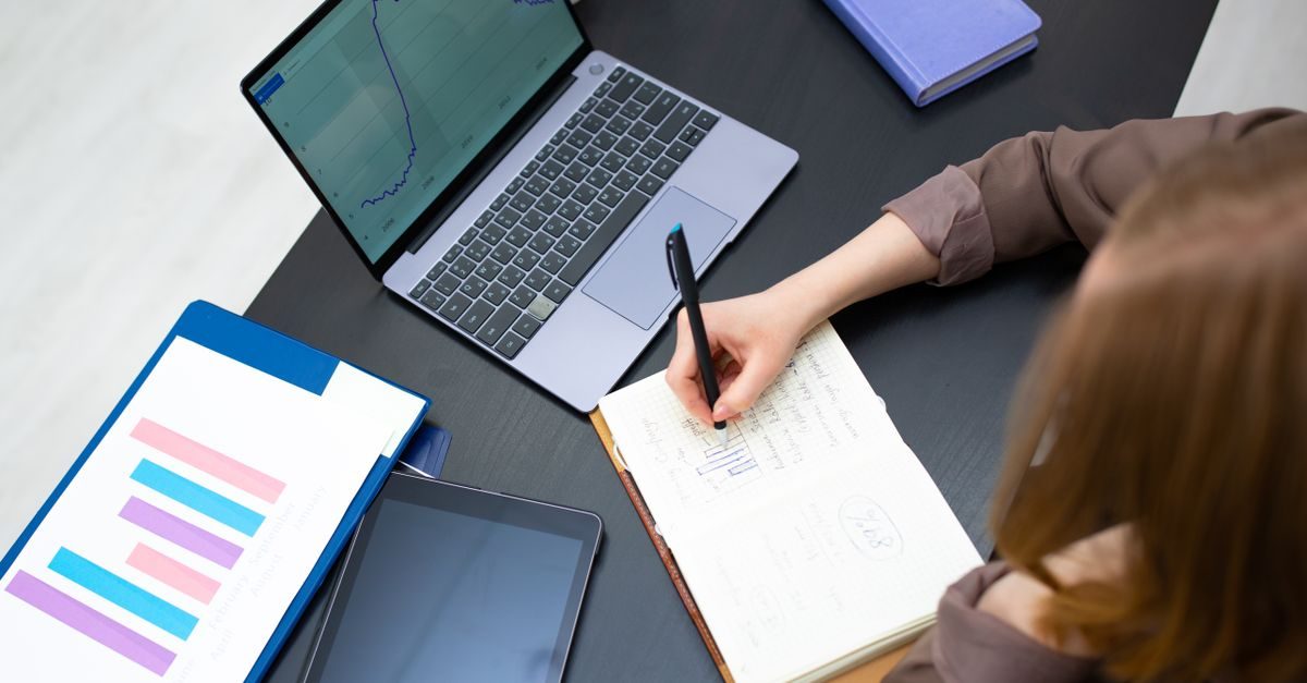 Una persona scrive su un quaderno accanto a un computer portatile con grafici e un tablet su una scrivania da ufficio