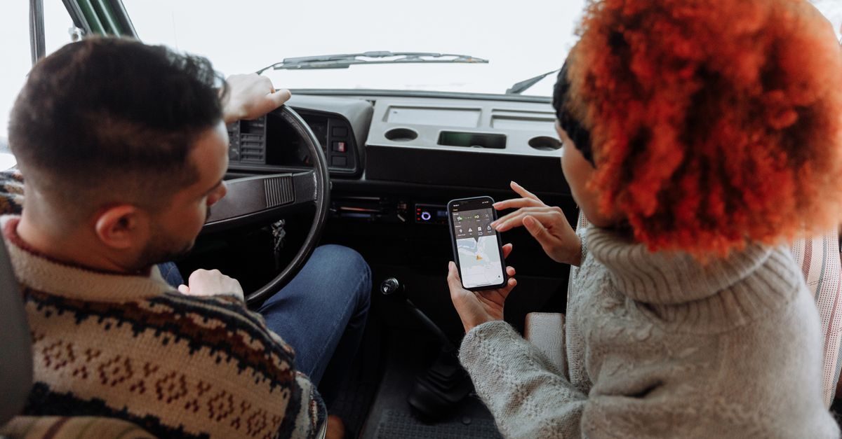 A man driving a van and a woman with red hair looking at a navigation app on a smartphone in the passenger seat