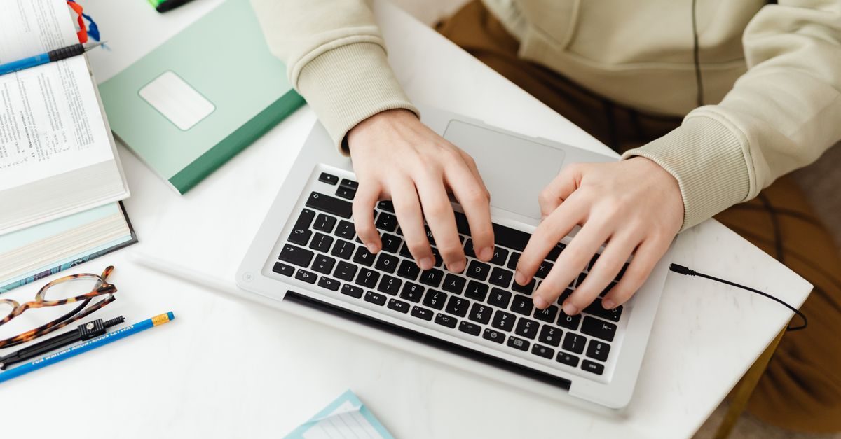 Hände tippen auf einer Laptop-Tastatur an einem Schreibtisch mit Notizbuch, Stiften und Brille in konzentrierter Atmosphäre