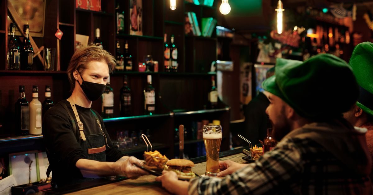 Un serveur masqué sert un burger et une bière à un client portant un chapeau vert dans un bar à l'ambiance tamisée