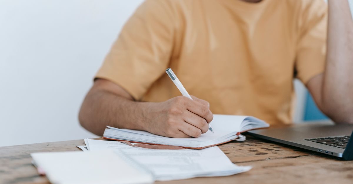 Persoon in een okergeel shirt schrijft met een pen in een notitieblok naast een laptop op een houten tafel