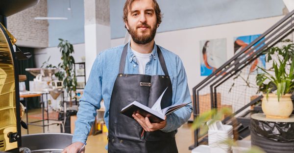 Hombre con barba y delantal negro sostiene un cuaderno en una cafetería con aspecto profesional y tranquilo