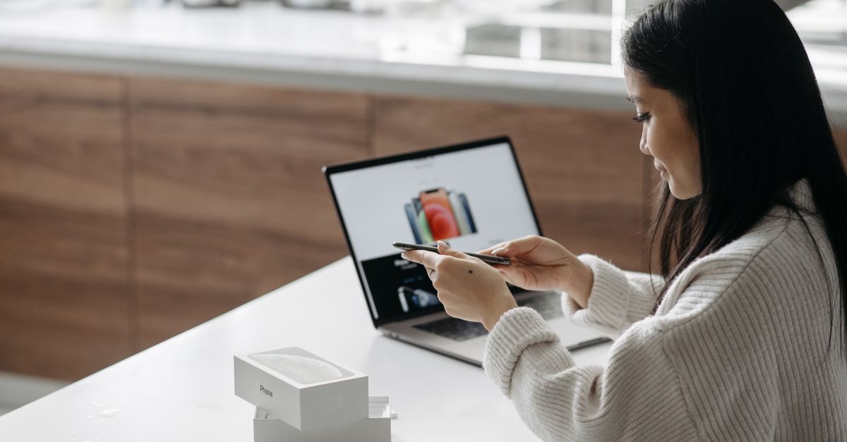 Eine junge Frau fotografiert mit ihrem Smartphone ein Produkt vor einem Laptop in einem hellen Büro
