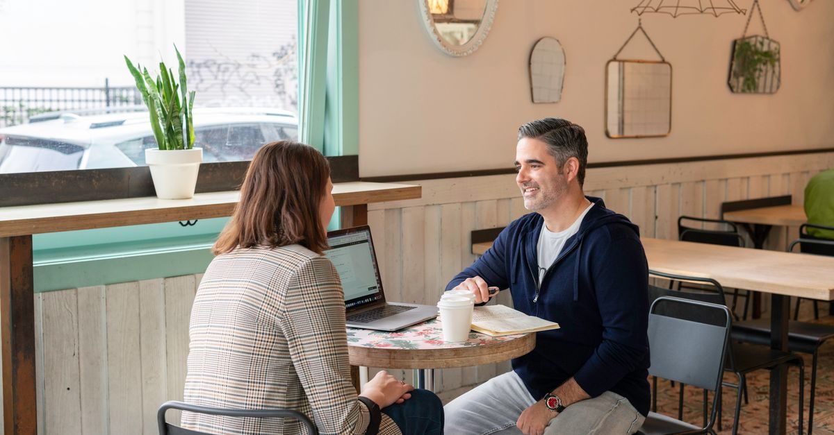 A man and woman sit at a cafe table with a laptop and coffee, engaged in a friendly professional conversation