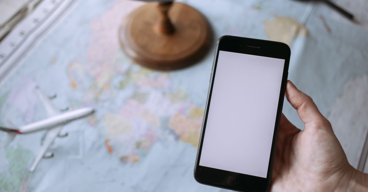 Mano che tiene uno smartphone con schermo bianco sopra una mappa geografica con un modellino di aereo e un mappamondo