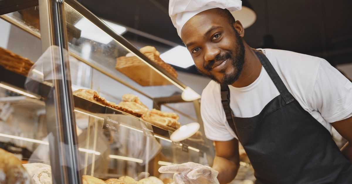 A smiling Black baker in a white chef hat and black apron stands behind a display case filled with fresh pastries