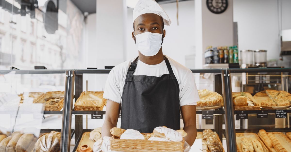 A masked baker in a white hat and black apron stands behind a display case filled with fresh bread in a bright bakery