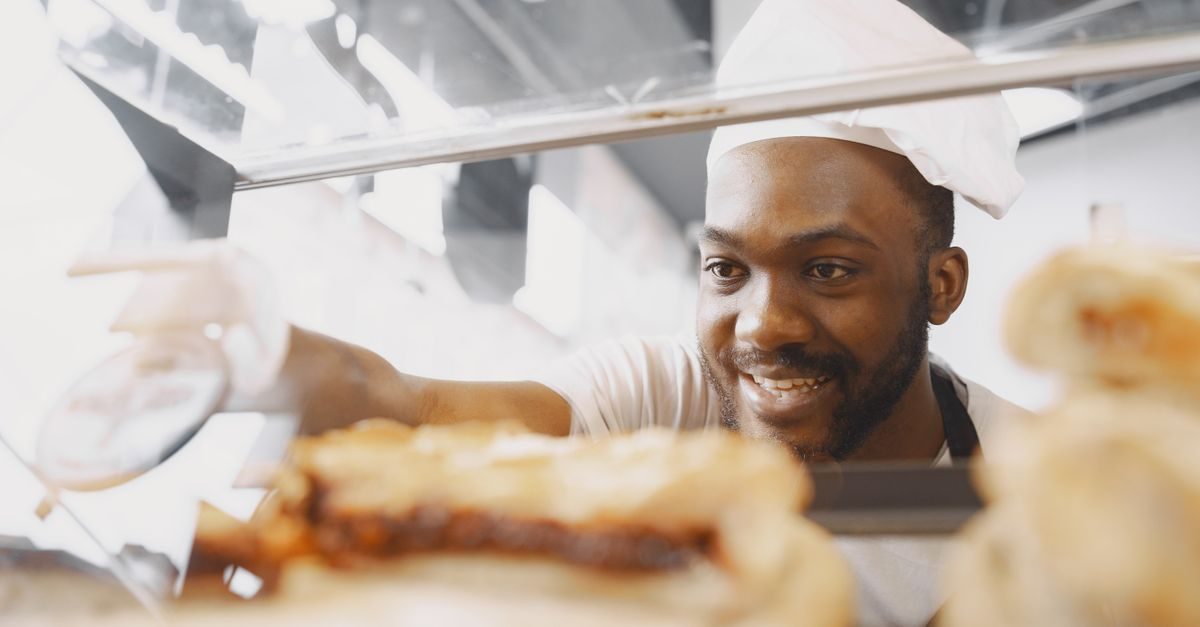 A smiling Black baker in a white hat reaches into a glass display case filled with pastries in a bright bakery shop