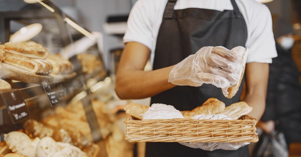 Bäcker mit schwarzer Schürze und Einweghandschuhen hält einen Korb mit frischem Gebäck in einer Bäckerei