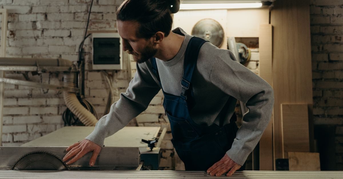Vakman in blauwe werkbroek werkt geconcentreerd aan een cirkelzaag in een werkplaats met bakstenen muur