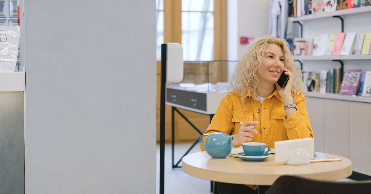 Une femme blonde en chemise jaune sourit en téléphonant à une table de café avec une théière et une tasse devant elle