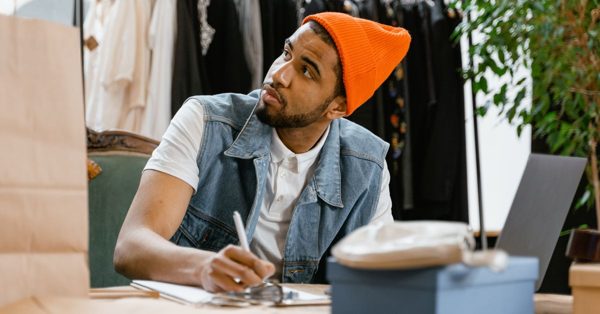 Un jeune homme en bonnet orange et gilet en jean prend des notes sur un carnet devant son ordinateur dans une boutique