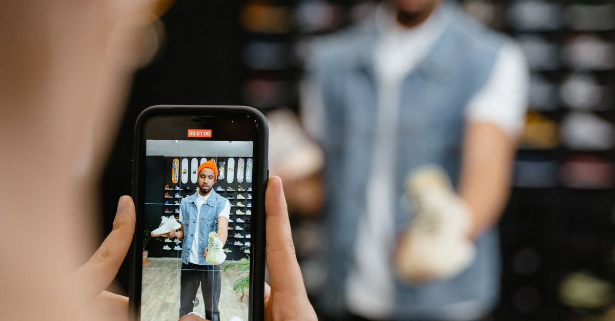 Persoon filmt met smartphone een man met muts die in een winkel sneakers toont aan de camera