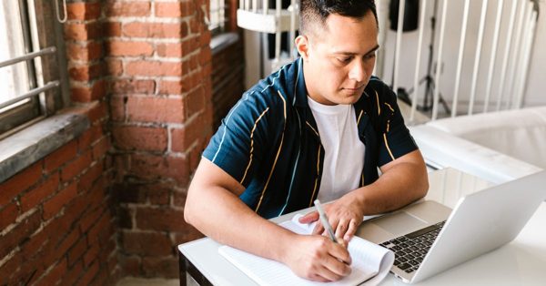 A focused man sits at a desk in a brick-walled office, writing in a notebook while looking at his open laptop