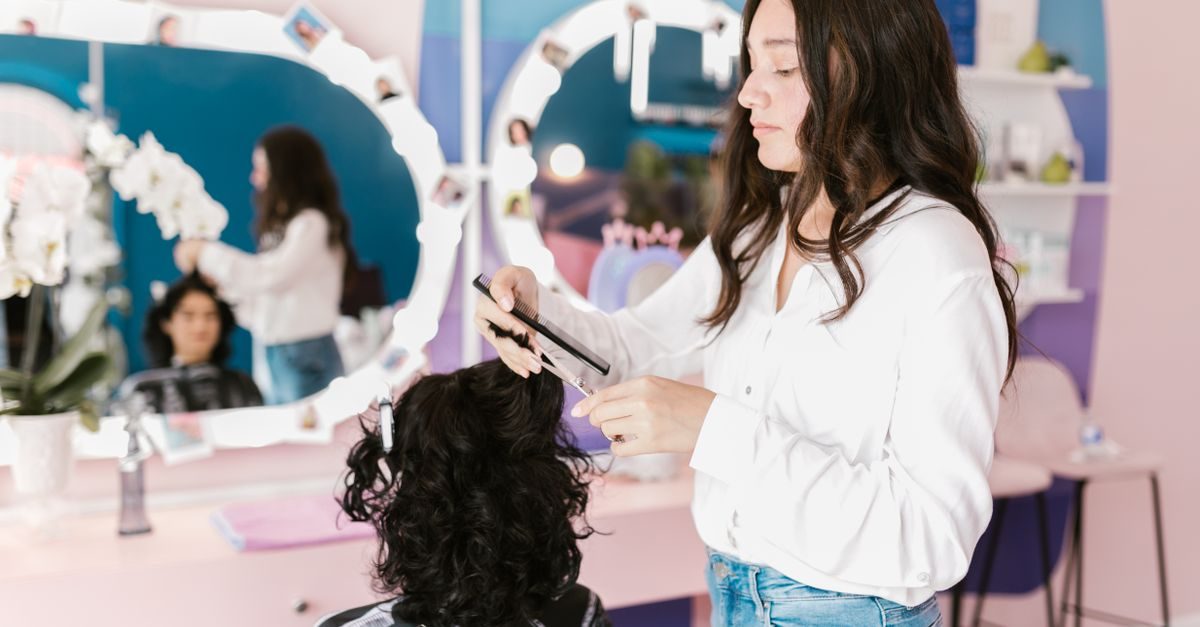 Une coiffeuse en chemise blanche coupe les cheveux d'une cliente dans un salon moderne aux tons roses et bleus