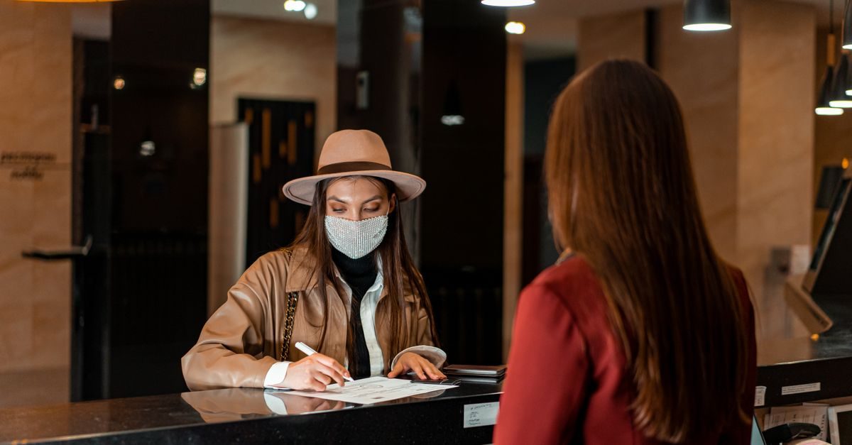 Vrouw met hoed en mondkapje vult een formulier in bij de hotelreceptie terwijl een medewerker toekijkt