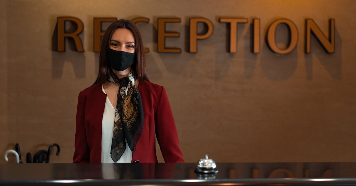 A smiling receptionist wearing a black face mask stands behind a hotel desk with a service bell in front of her