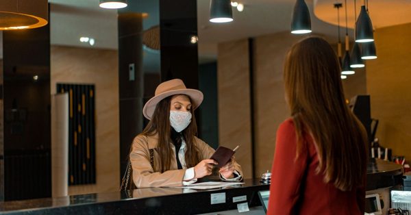 A woman in a hat and face mask checks into a hotel, handing a passport to a receptionist at a modern front desk